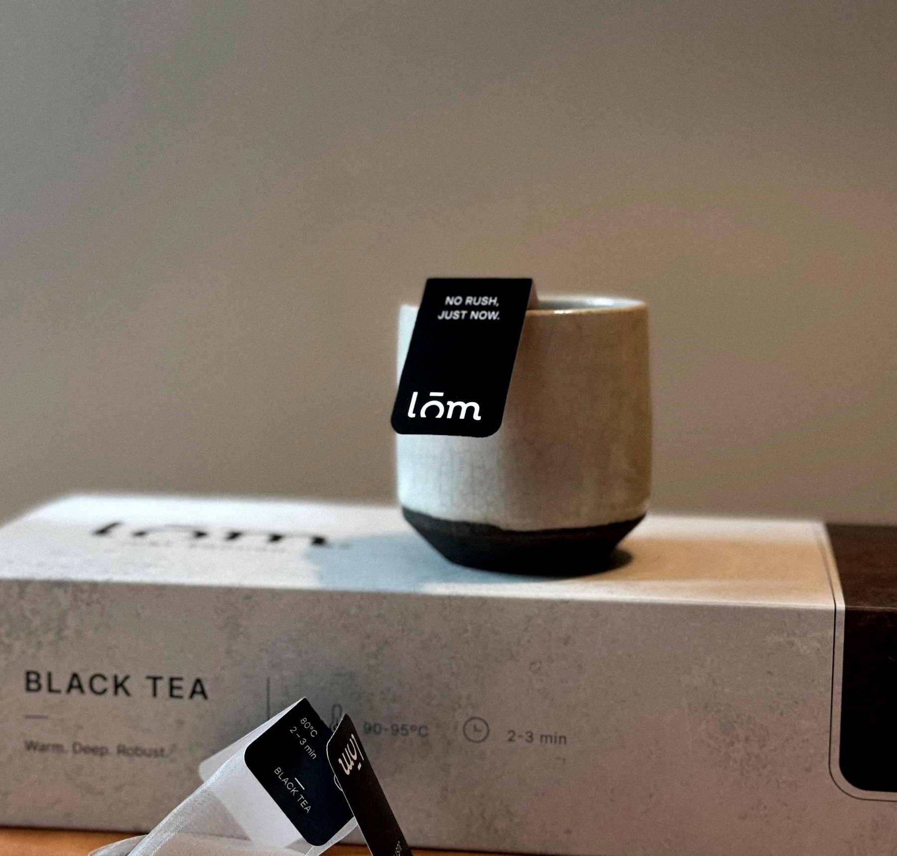 Taza de cerámica con la bolsita de té LŌM anclada al borde, junto a la caja de English Breakfast y un sobre individual sobre superficie neutra